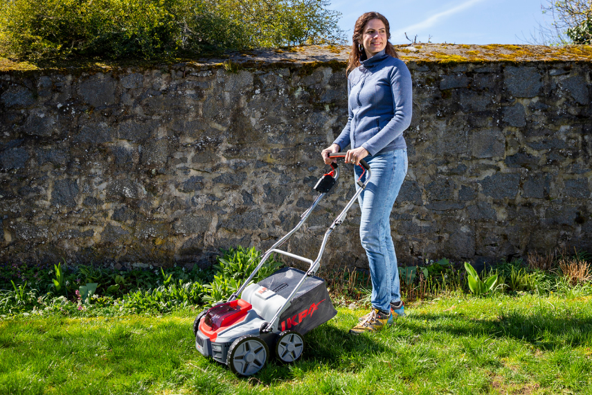 Eine Frau steht mit dem BOSCH Akku Vertikutierer IB-SR 36 auf einem grünen Rasen vor einer Gartenmauer.